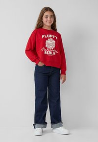 Rode sweatshirt met de grafische tekst "FLUFFY Pancakes BERLIN", met een relaxte pasvorm en brede mouwen, gecombineerd met donkere denim jeans en witte sneakers.