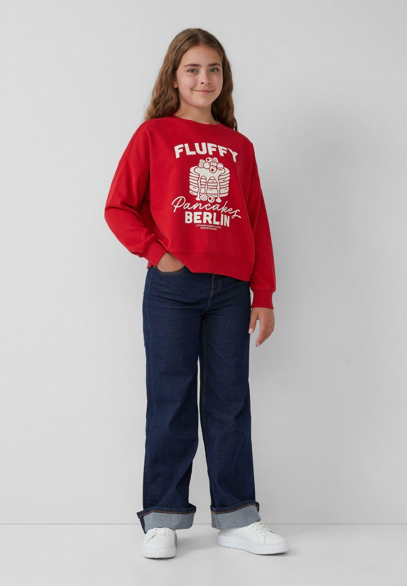 Rode sweatshirt met de grafische tekst "FLUFFY Pancakes BERLIN", met een relaxte pasvorm en brede mouwen, gecombineerd met donkere denim jeans en witte sneakers.