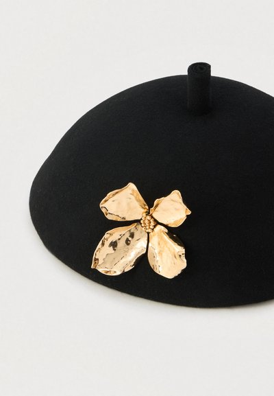 Mustast feltmütsist, millel on ümar kuju ja reljeefse tekstuuriga kuldne lillekujundus. Lillel on kroonlehe detailid ja keskel pärlikujuline mustriga aksent.