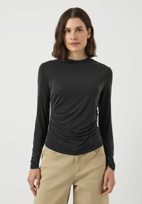 Svart långärmad topp i ett mjukt, draperat tyg med rund halsringning och samlade detaljer vid midjan, ihopparad med beige byxor.