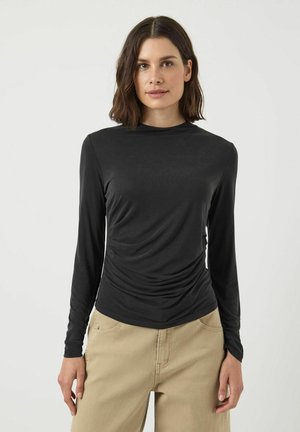 Maglietta nera a maniche lunghe in un morbido tessuto drappeggiato, con scollatura rotonda e dettagli arricciati in vita, abbinata a pantaloni beige.