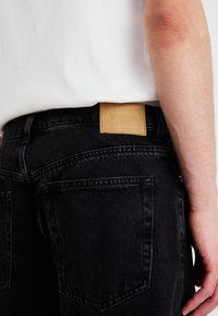 Jeans negri din denim au un eticheta din piele cu text embosat, cusături ușor vizibile și o vedere de aproape a unui buzunar din spate.