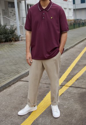 Man die een bordeauxrode poloshirt, beige broek met omslagen en witte sneakers draagt, staand op straat naast een gele lijn.