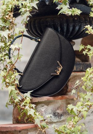 Černá kabelka s texturovaným povrchem, trojúhelníkového tvaru, s akcenty ze zlaceného kovu a krátkým uchem, umístěná na kamenném pozadí s břečťanem.