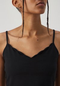 Haut noir sans manches avec un design en col en V à bords festonnés, fabriqué en tissu doux, mettant en valeur une texture lisse et une coupe ajustée.