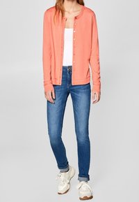 Korálový kardigán s knoflíky, nošený přes bílý tílko, spárovaný s modrými džíny a bílými teniskami s hnědými akcenty.