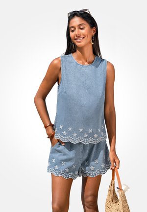 Femme portant un top en jean sans manches et un short assorti avec des bords brodés blancs, tenant un sac à main tissé avec des poignées marron.