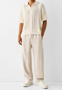 Polo de punto color crema con un patrón vertical texturizado, combinado con pantalones beige holgados y zapatillas blancas. Diseño de cuello simple.
