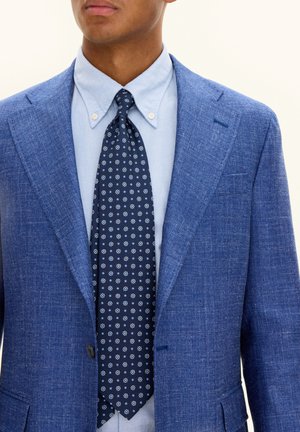 Blazer blu con una finitura testurizzata, camicia azzurra e cravatta blu navy con motivi floreali bianchi e azzurri.