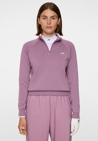 Eine Frau trägt einen mauvefarbenen Reißverschluss-Pullover und passende Hosen, während sie einen weißen Golfhandschuh an ihrer rechten Hand gegen einen neutralen Hintergrund trägt.