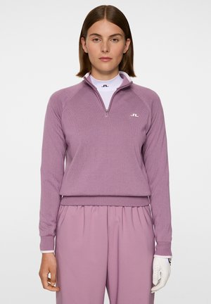 Nainen, jolla on päällään mauvenvärinen vetoketjullinen neule ja siihen matching-housut, sekä valkoinen golf-käsine oikeassa kädessään tasaisella taustalla.