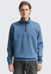 Blauer Pullover mit hohem Kragen, halbem Reißverschluss und langen Ärmeln. Der Stoff wirkt weich und hat eine glatte Textur.