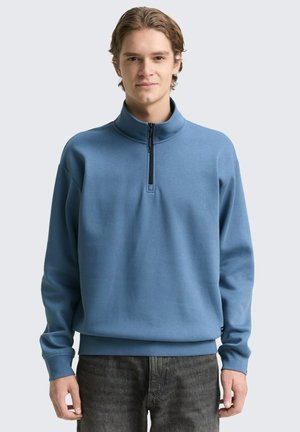 Blauer Pullover mit hohem Kragen, halbem Reißverschluss und langen Ärmeln. Der Stoff wirkt weich und hat eine glatte Textur.