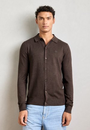 Brown button-up cardigan with a collar, knit texture, ribbed cuffs, and hem, worn over light blue jeans. Subtle logo detail on chest.