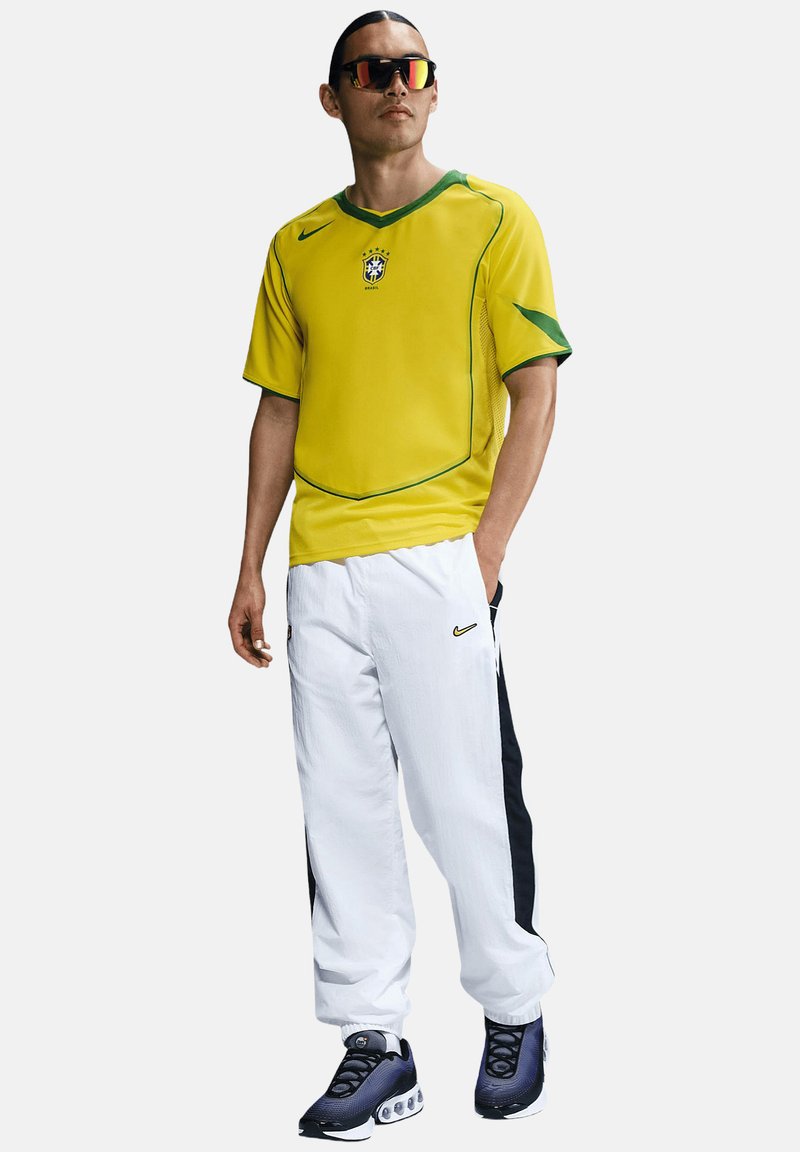 Maglia da calcio Nike gialla con dettagli verdi, abbinata a pantaloni da allenamento bianchi con strisce nere laterali. Scarpe sportive nere e argento.