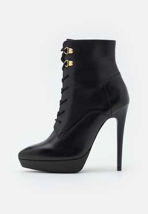 Bottine en cuir noir avec un bout pointu, un talon haut stilettos, un devant lacé et des accents en métal doré. Texture lisse.
