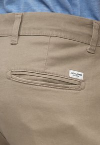 Gros plan sur la poche arrière d'un pantalon beige avec l'étiquette de la marque Jack & Jones et une partie d'une chemise bleue visible au-dessus de la ceinture.