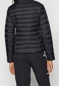 Sort pufferjakke med en quiltet design, prydet med vandrette syninger, en høj krave og en tætsiddende pasform. Sammen med sorte bukser.