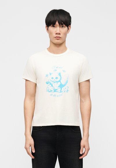 Jeune homme portant un t-shirt crème à manches courtes avec un motif de chat bleu et un texte, associé à un pantalon noir, debout devant un fond clair uni.