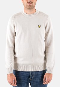 Pull beige clair avec col et ourlet côtel�és, présentant une texture subtile et un petit logo brodé jaune sur la poitrine.