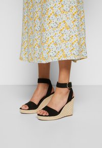Black wedge sandals with a textured espadrille sole, adjustable ankle strap, and open toe design. Suitable for casual wear.