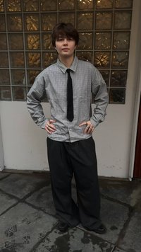 Άτομο με κοντά σκούρα μαλλιά στέκεται με τα χέρια στη μέση φορώντας πουκάμισο με καρό σχέδιο, μαύρη γραβάτα, άνετο μαύρο παντελόνι και δαχτυλίδια, μπροστά από γυάλινες πλάκες.