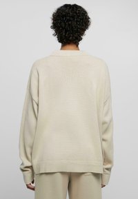 Jasno-beżowy oversize sweter z prążkowanym dołem i mankietami, z dzianinową fakturą i klasycznym kołnierzykiem. Widok z tyłu.