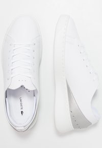 Zapatillas blancas de material sintético liso, con un diseño minimalista, cordones planos y un detalle plateado en el talón.