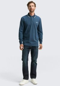 Langarm-Polohemd in dunkelblau mit weißem Kragen und Akzenten, ausgestattet mit einer Drei-Knopf-Leiste. Kombiniert mit dunklen Denim-Jeans und weißen Turnschuhen.