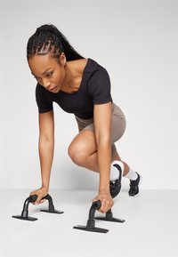 Black exercise push-up bars on a light surface. Person in a fitted black top and beige shorts prepares to use them, with a focused stance.