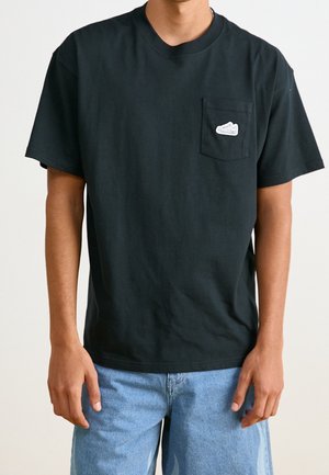 Jong persoon draagt een effen zwart T-shirt met korte mouwen en een klein wit sneakers-logo op het linkerborstzakje, gecombineerd met lichtblauwe spijkerbroek.