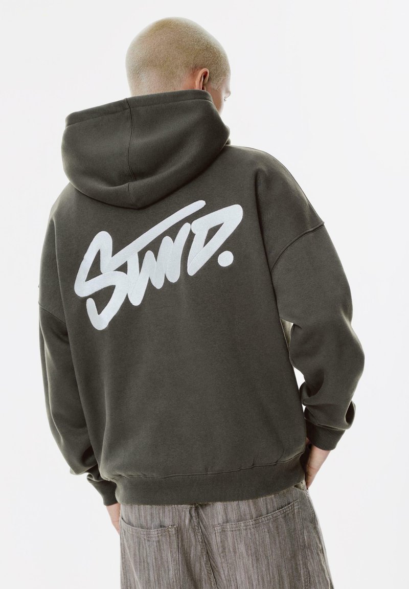 Felpa con cappuccio verde scuro con un grande logo stilizzato bianco sulla schiena. Presenta un cappuccio, polsini a coste e una vestibilità comoda. Tessuto morbido.