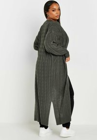 Lång grön stickad kofta med flätstickat mönster, ribbade ärmavslut och sidor. Bärs över svarta byxor och vita sneakers.