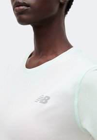 Κοντινό πλάνο ενός ατόμου που φοράει ένα ανοιχτό πράσινο μπλουζάκι New Balance με στρογγυλή λαιμόκοψη, ορατό από τους ώμους μέχρι το πηγούνι.