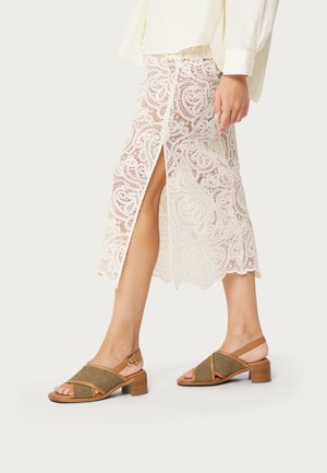 Mujer con una falda midi de encaje crema con abertura frontal y sandalias de tacón grueso color cuero con tiras en el tobillo.