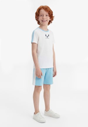 Enfant roux souriant portant une tenue de sport Messi blanche et bleu clair avec des baskets sur un fond blanc uni.