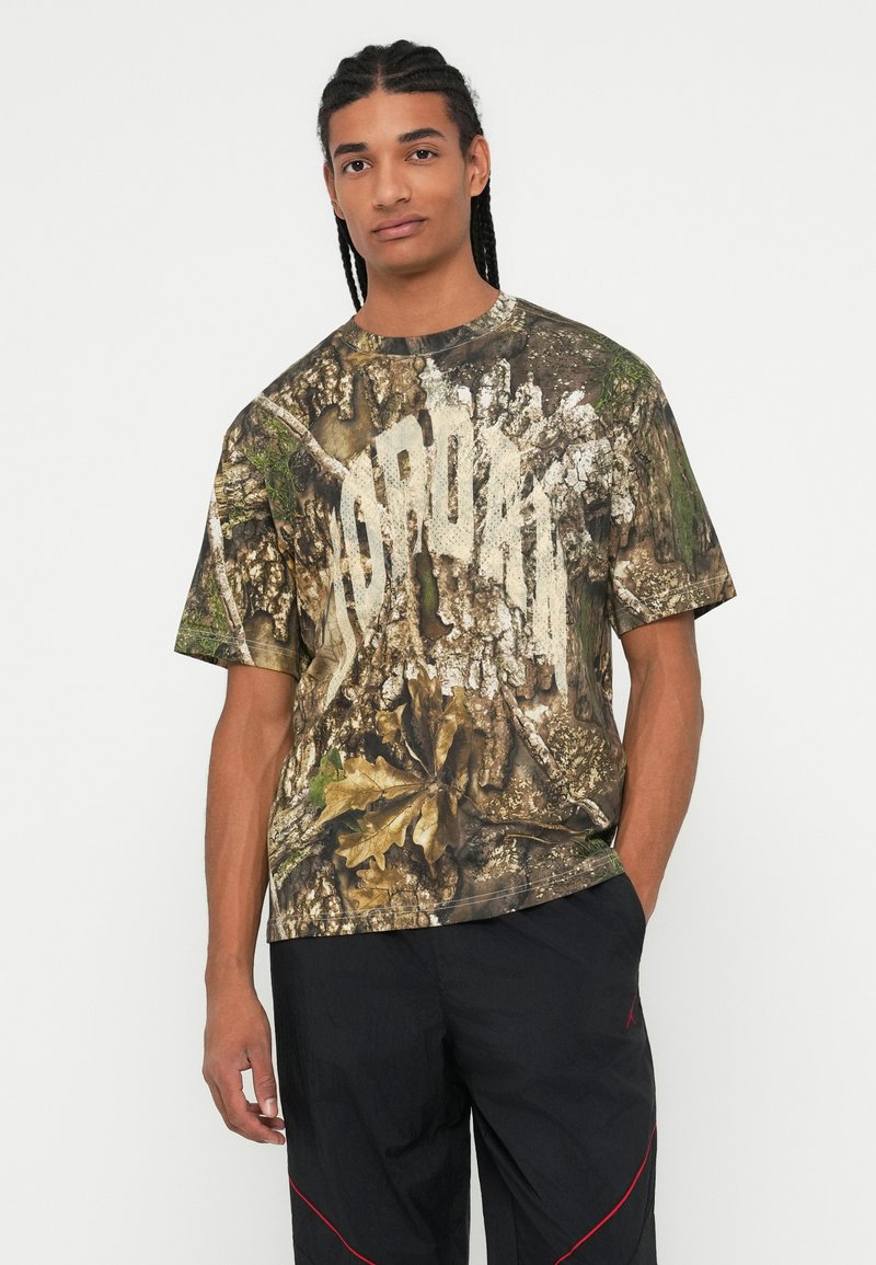 Homme avec des cheveux tressés portant un t-shirt à imprimé camouflage et un pantalon noir avec des lignes rouges, se tenant avec une main dans la poche sur un fond blanc.