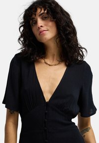 Μαύρο φόρεμα με κοντά μανίκια σε στυλ φτερού, V-λαιμόκοψη και υφασμάτινη υφή. Διαθέτει σειρά από κουμπιά καλυμμένα με ύφασμα στο μπροστινό μέρος.