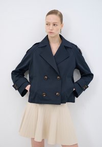 Mörkblå dubbelknäppt jacka i slät tyg, med stora bruna knappar, axelklaffar och breda slag. Stylingen kompletteras med en beige veckad kjol.