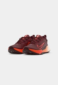 Zapatillas deportivas rojas y naranjas con parte superior de malla, suela texturizada y cordones. Se pueden ver detalles en negro y el logotipo en el lateral.