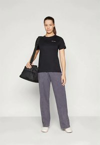 Ung kvinne står med en svart treningsbag over skulderen, iført en svart t-skjorte, løse grå bukser og hvite joggesko på en ensfarget bakgrunn.