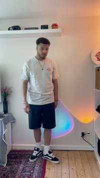 Camiseta gris de manga corta con logo rosa, pantalones cortos negros y zapatillas negras con acentos blancos. Suelo de madera y fondo de luces coloridas.