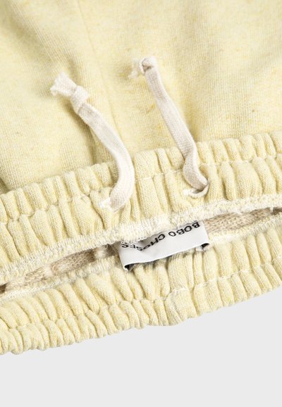 Gros plan sur une taille élastique beige avec cordons et une étiquette en tissu blanc partiellement visible sur un vêtement à texture douce.