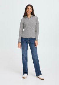 Pilkas raštuotas megztinis su užtrauktuku ant apykaklės, derinamas su mėlynomis džinsinėmis platėjančiomis kelnėmis ir baltais sportbačiais, esant neutraliam fone.