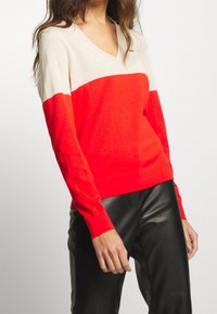 Svetr s výstřihem do V, který má krémovou horní část a živě červenou dolní část, vyrobený z měkkého pleteného materiálu; přiléhavé rukávy s žebrovanými manžetami.