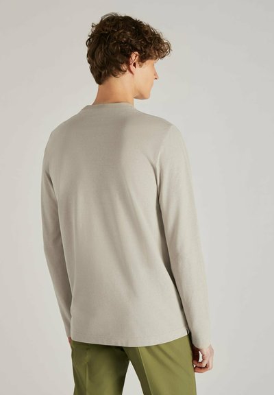 Camisa de manga larga color beige claro, con cuello redondo, textura suave, ajuste relajado, mostrada desde atrás con pantalones verdes oliva visibles en la cintura.