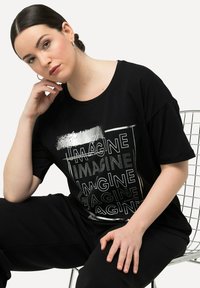 Camiseta negra con el texto en blanco "IMAGINE" en diferentes fuentes y acentos metálicos. Ligeramente holgada con un escote redondeado.