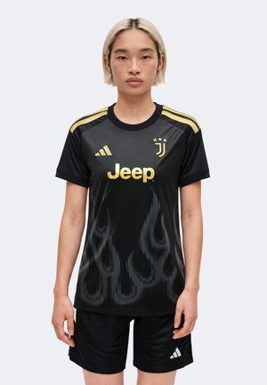 Personne aux cheveux blonds courts portant un maillot de football noir et or de la Juventus et un short Adidas noir, debout devant un fond uni.