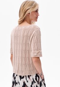 Jasnobeżowy sweter z dzianiny o fakturowanym wzorze, z krótkimi, podwiniętymi rękawami i luźnym krojem, noszony na wzorzystą spódnicę.