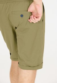 Olijfgroene shorts van lichtgewicht stof, met opgerolde zoom en een achterzak met een blauwe knoopdetail.